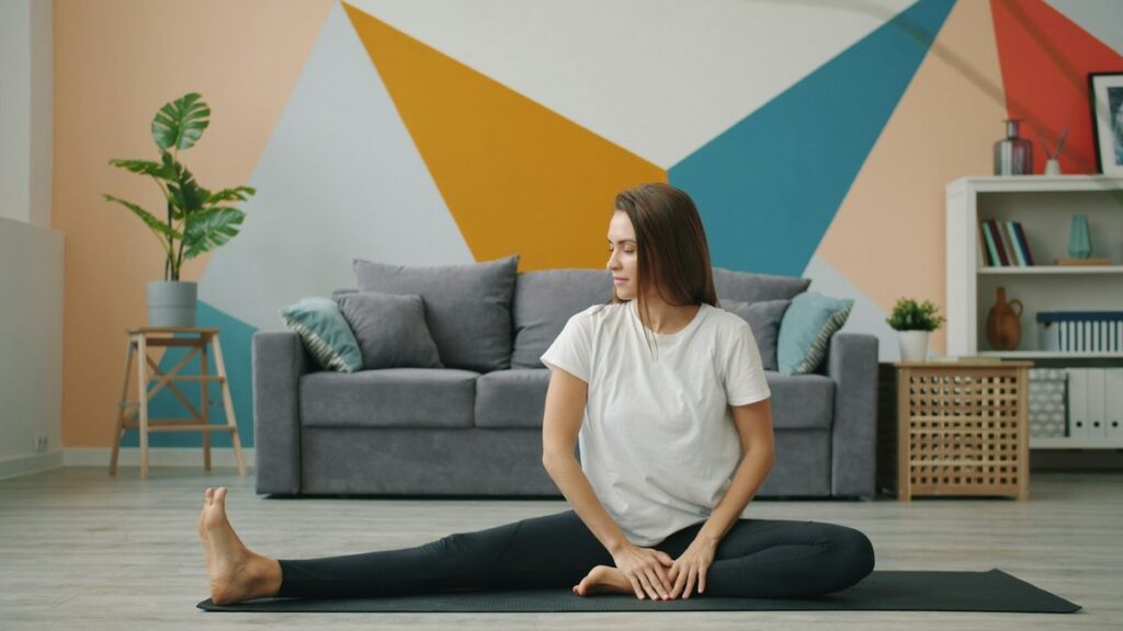 Mujer sentada en una esterilla haciendo estiramientos después de entrenar en casa, en el salón